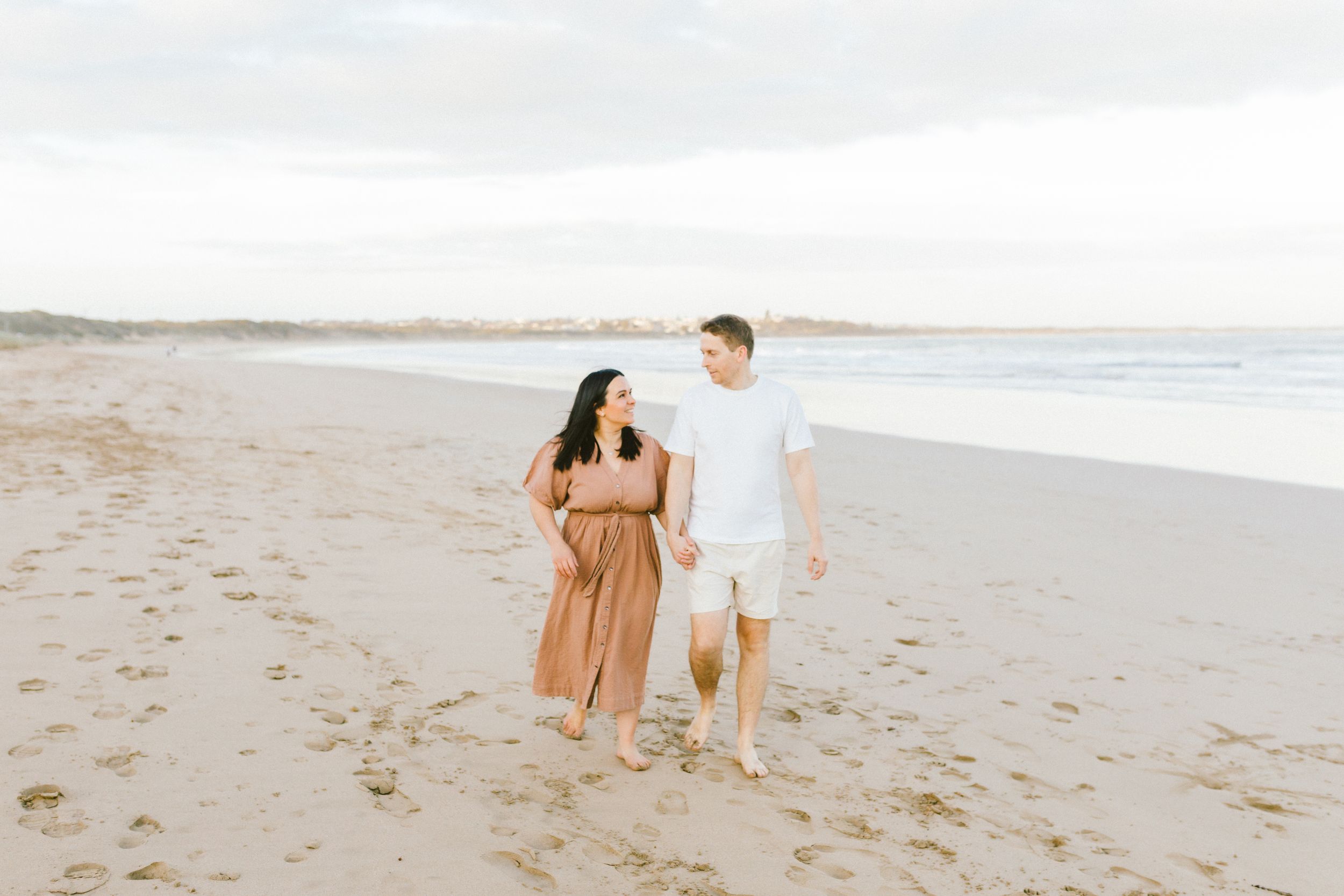 A couple in brown outfits walks along a sandy beach during sunset creating romantic silhouettes.