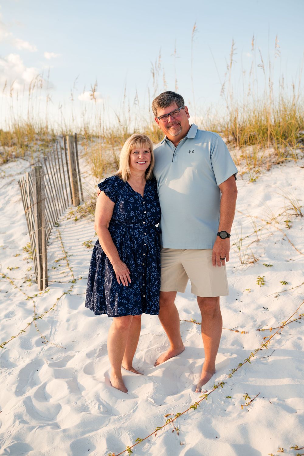 A couple stands together on a white sandy beach with sea oats and wooden fencing in the background during sunset.