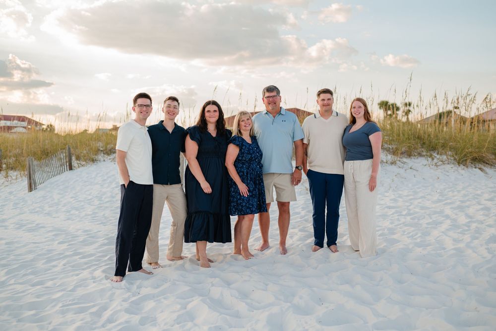 A group of people wearing coordinated blue and white outfits pose together on a white sandy beach at sunset.