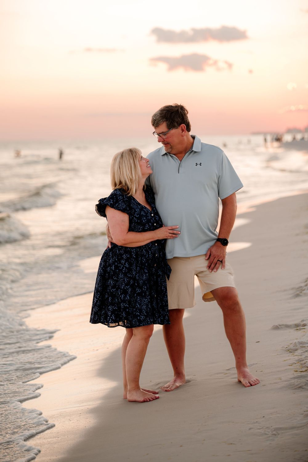 A couple embraces while walking along a sandy beach at sunset with gentle waves rolling in along the shore.