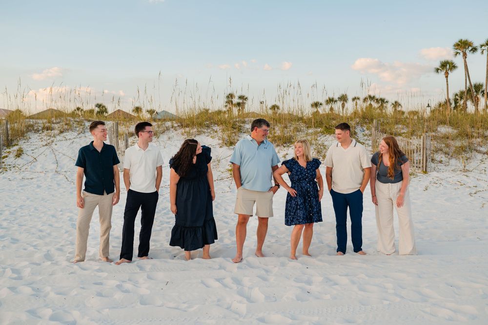A group of people stand together on a white sandy beach with palm trees visible in the background during sunset.