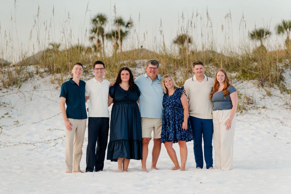 Group of people standing together on a white sandy beach at sunset wearing coordinated blue and white outfits.