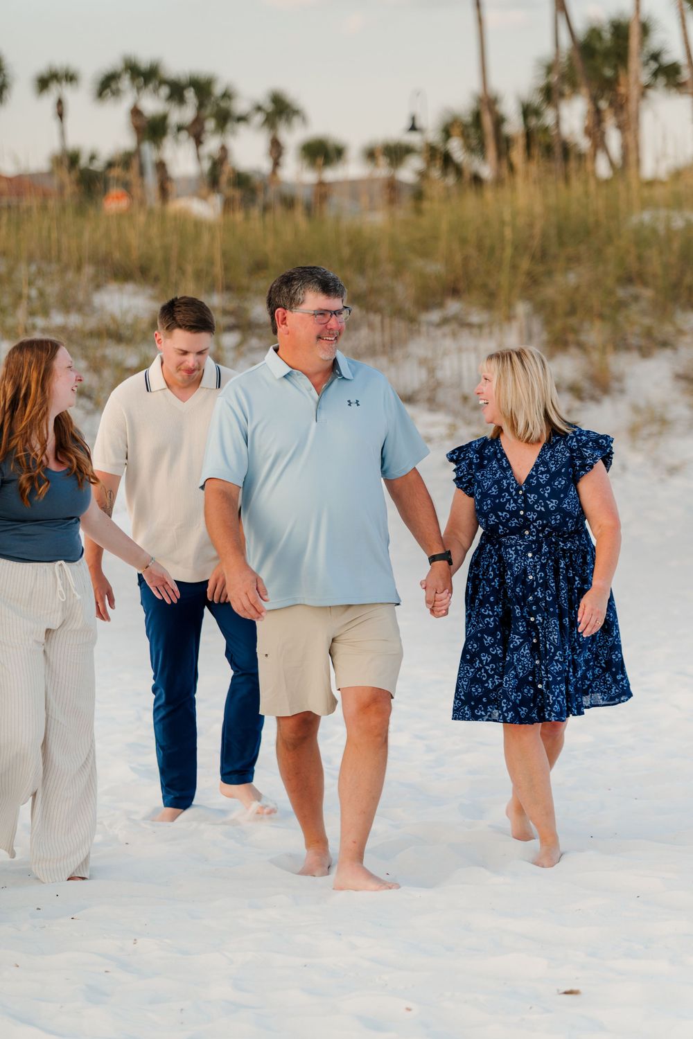 Family group walking together on white sandy beach with palm trees in background wearing coordinated blue and white outfits.