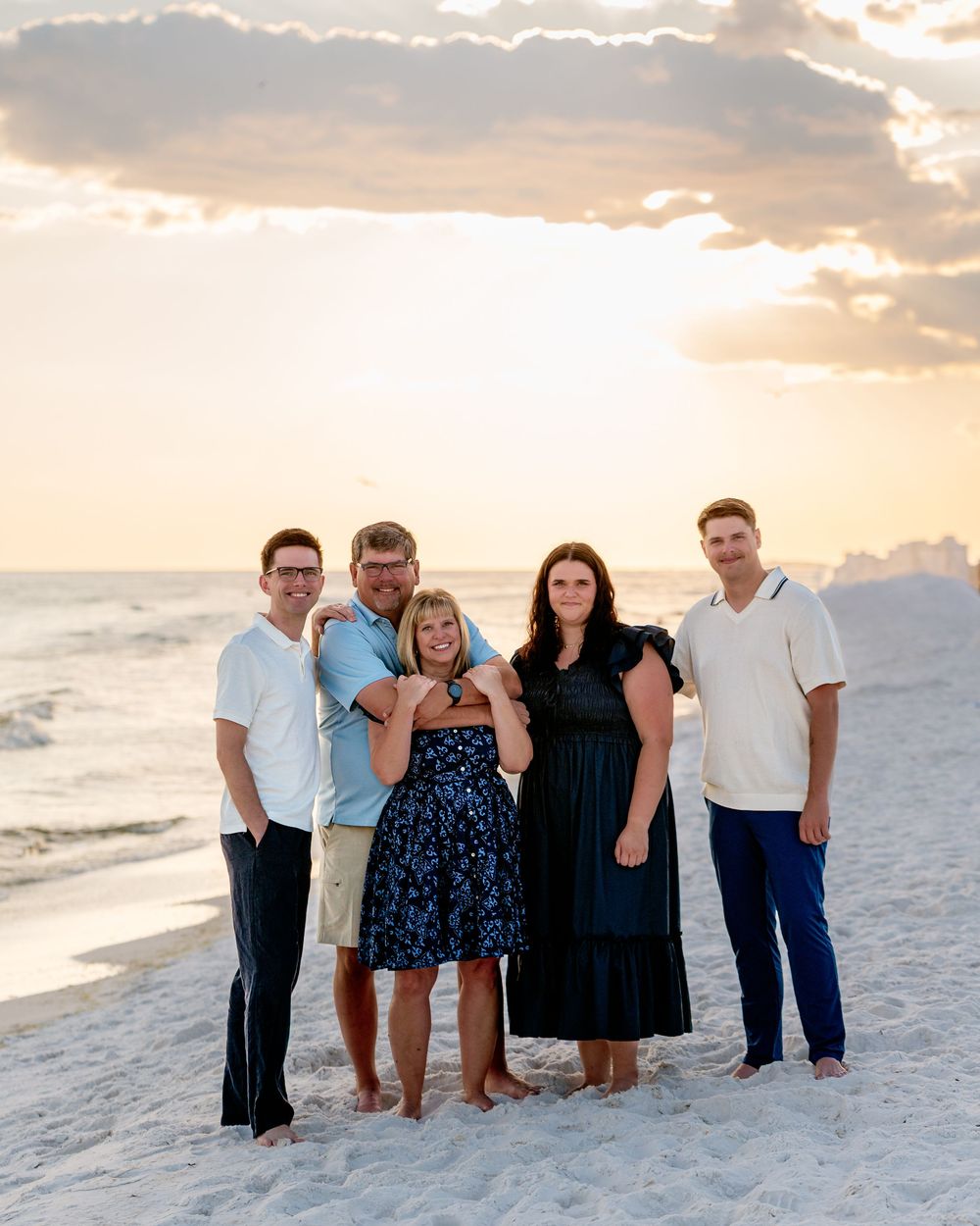 A group of people stand together on a white sandy beach at sunset with waves and clouds in the background.