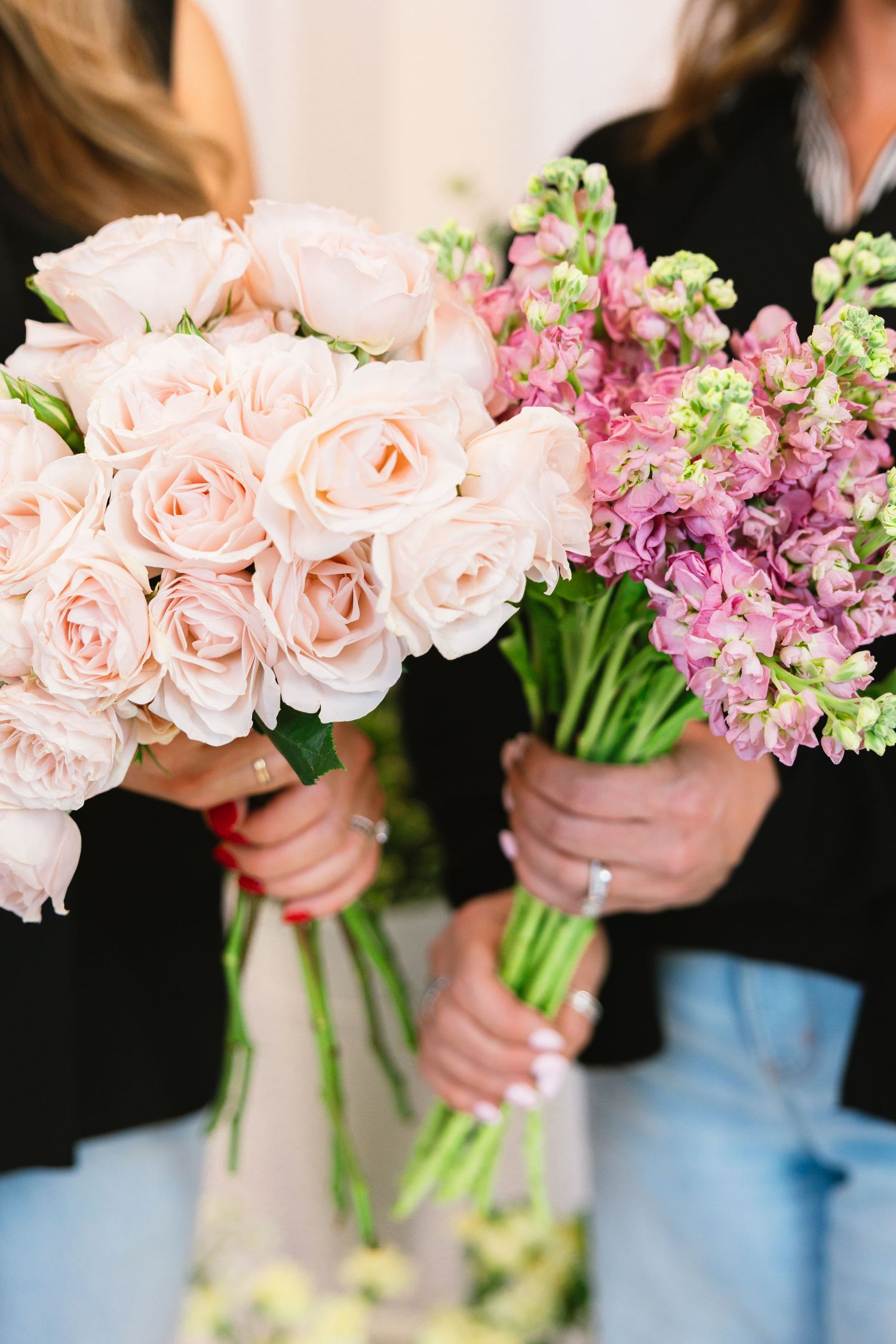 Two women in floral dresses stand beside a white tiered display shelf filled with colorful potted flowers and plants.