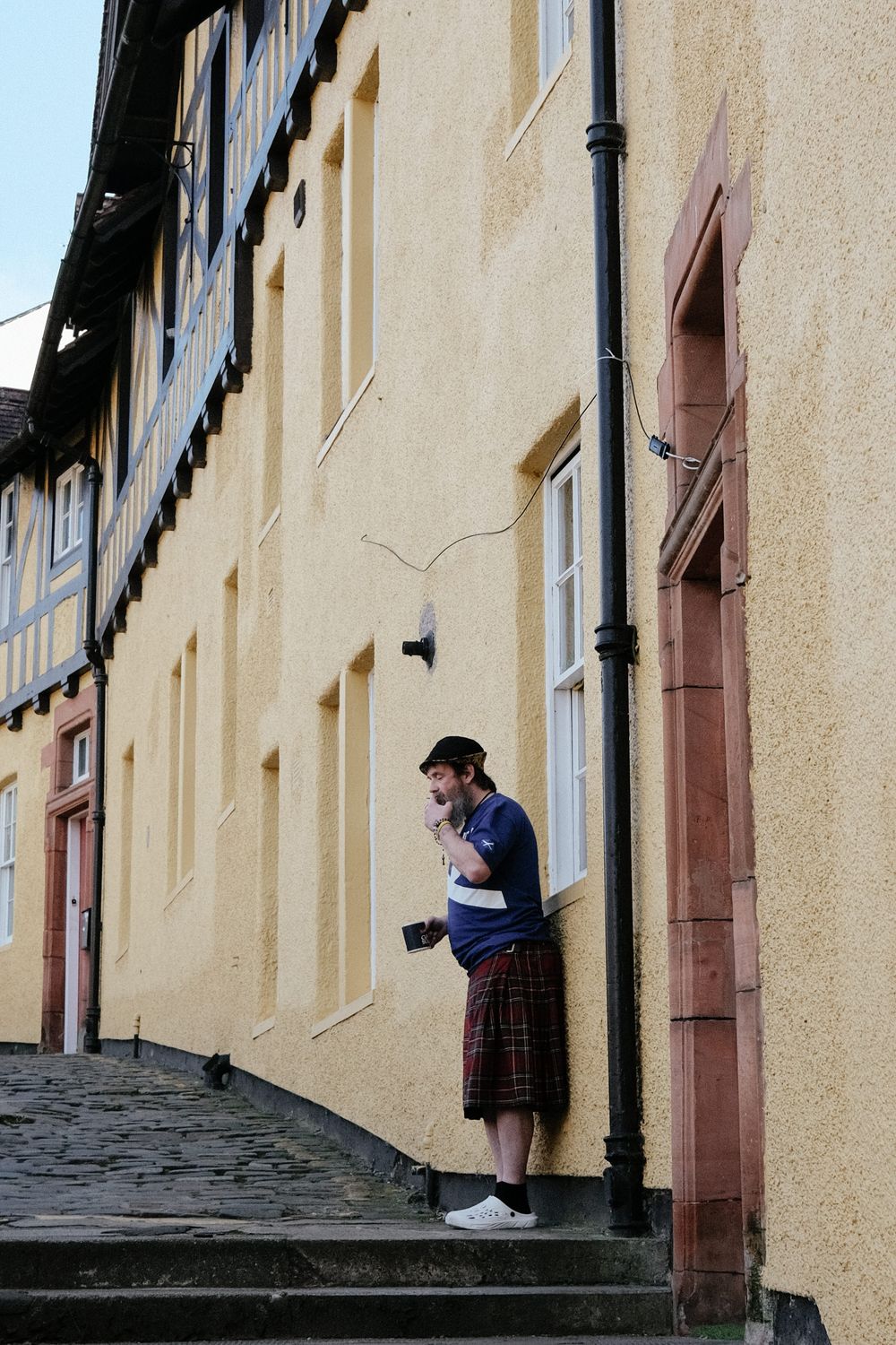 Someone wearing a plaid skirt and dark top stands on stone steps against a yellow building wall in a historic town.