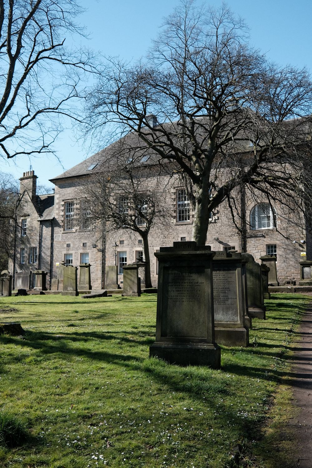 Stone gravestones scattered across a grassy cemetery with historic buildings in the background.