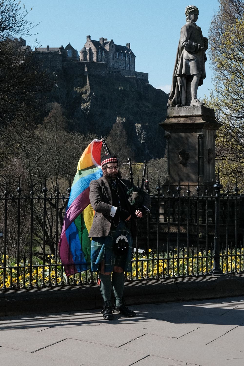 People with colorful pride flags gather near statues and iron railings with Edinburgh Castle in background.