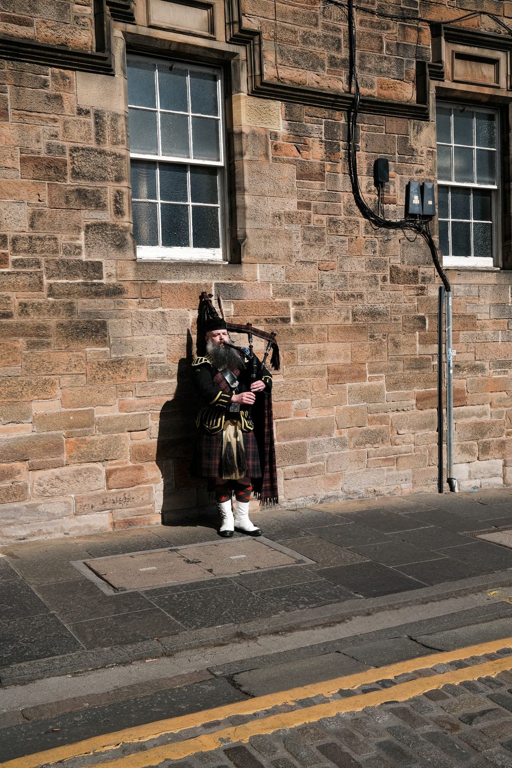 A bagpiper in traditional Scottish attire plays music on a stone-walled street in Edinburgh.