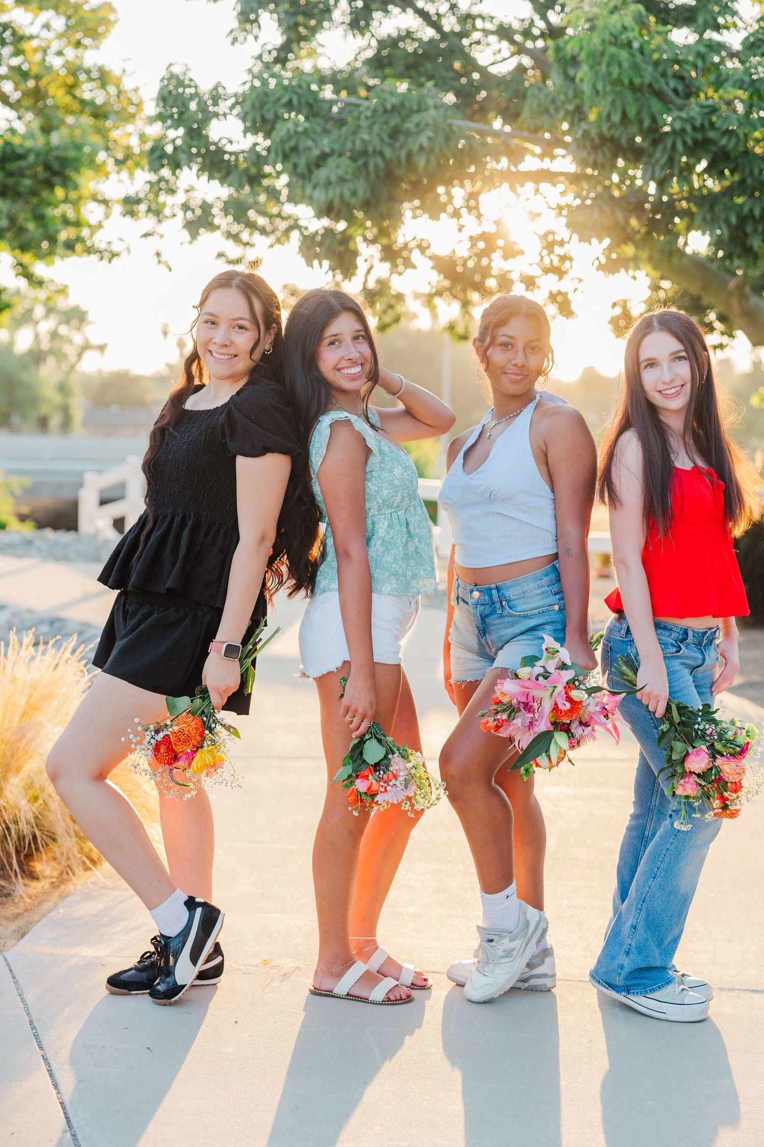 Four friends pose together at sunset holding colorful flower bouquets while wearing casual summer outfits.