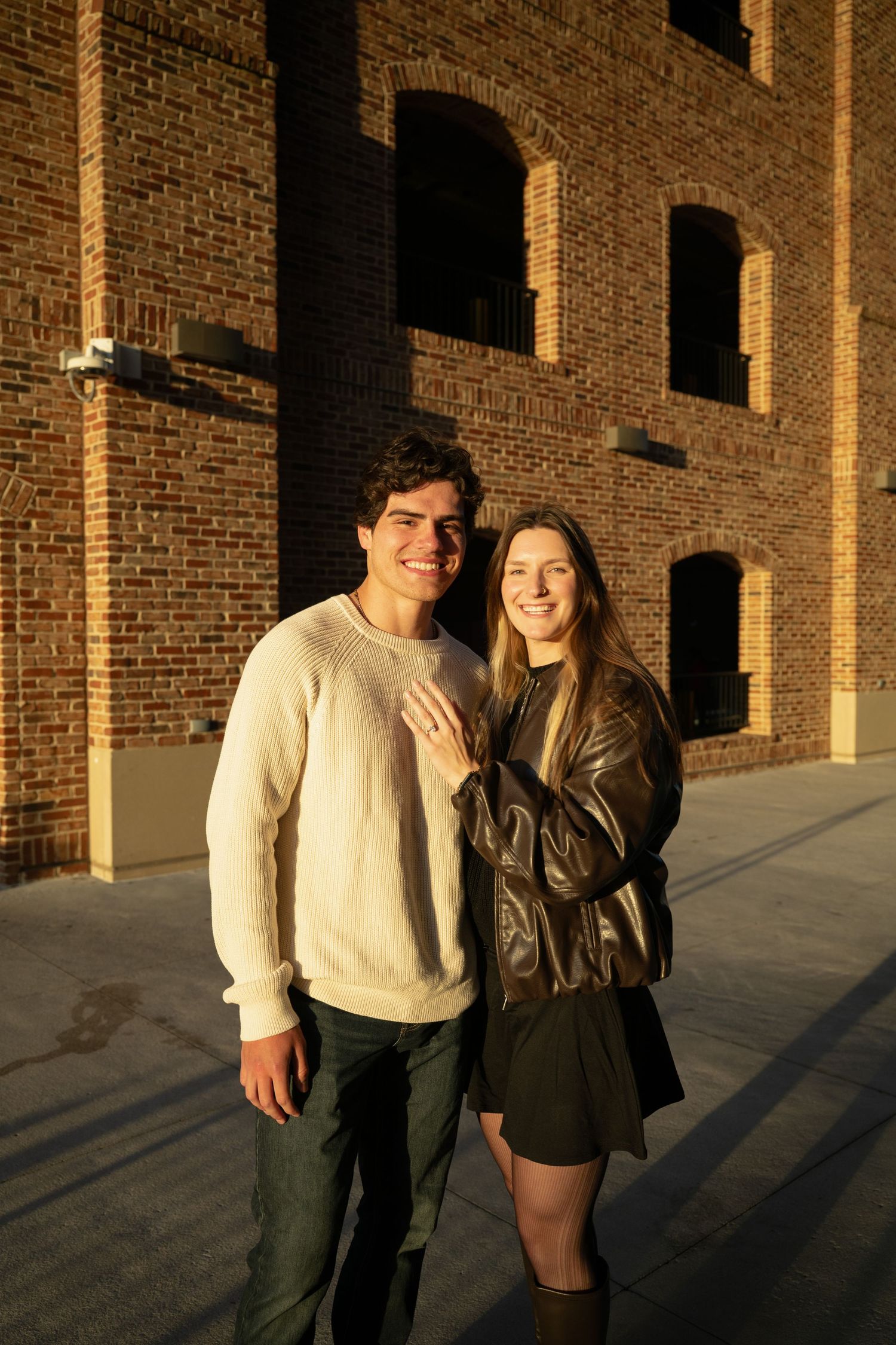 A couple stands together against a brick wall in warm sunlight casting dramatic shadows.