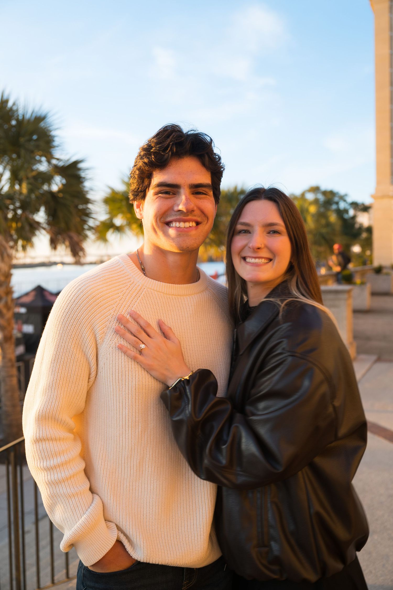 A couple smiles warmly at the camera while embracing outdoors, with one wearing a cream sweater and the other in a black jacket.