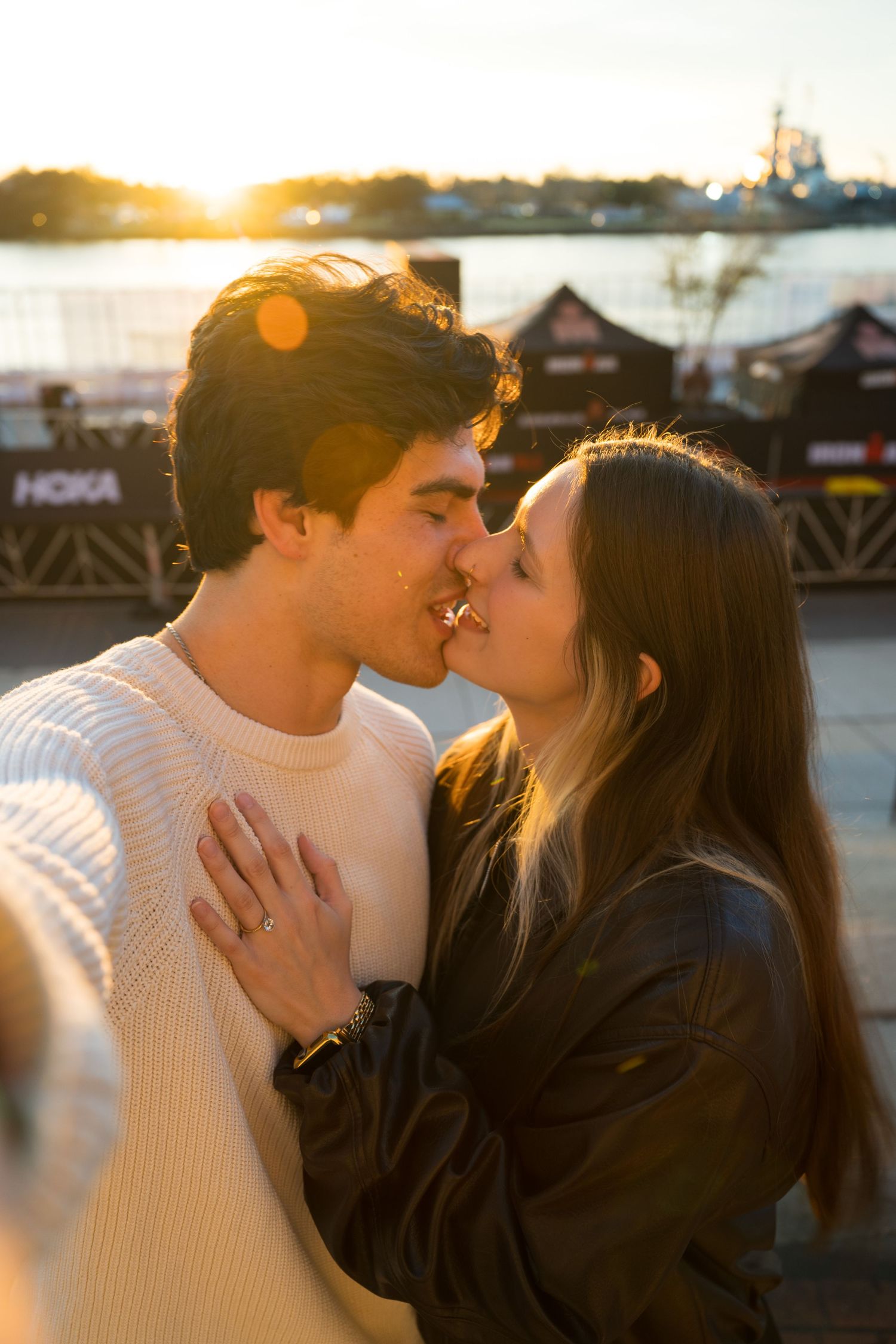 A romantic couple shares a kiss at sunset with a waterfront backdrop, dressed in contrasting cream and black outfits.