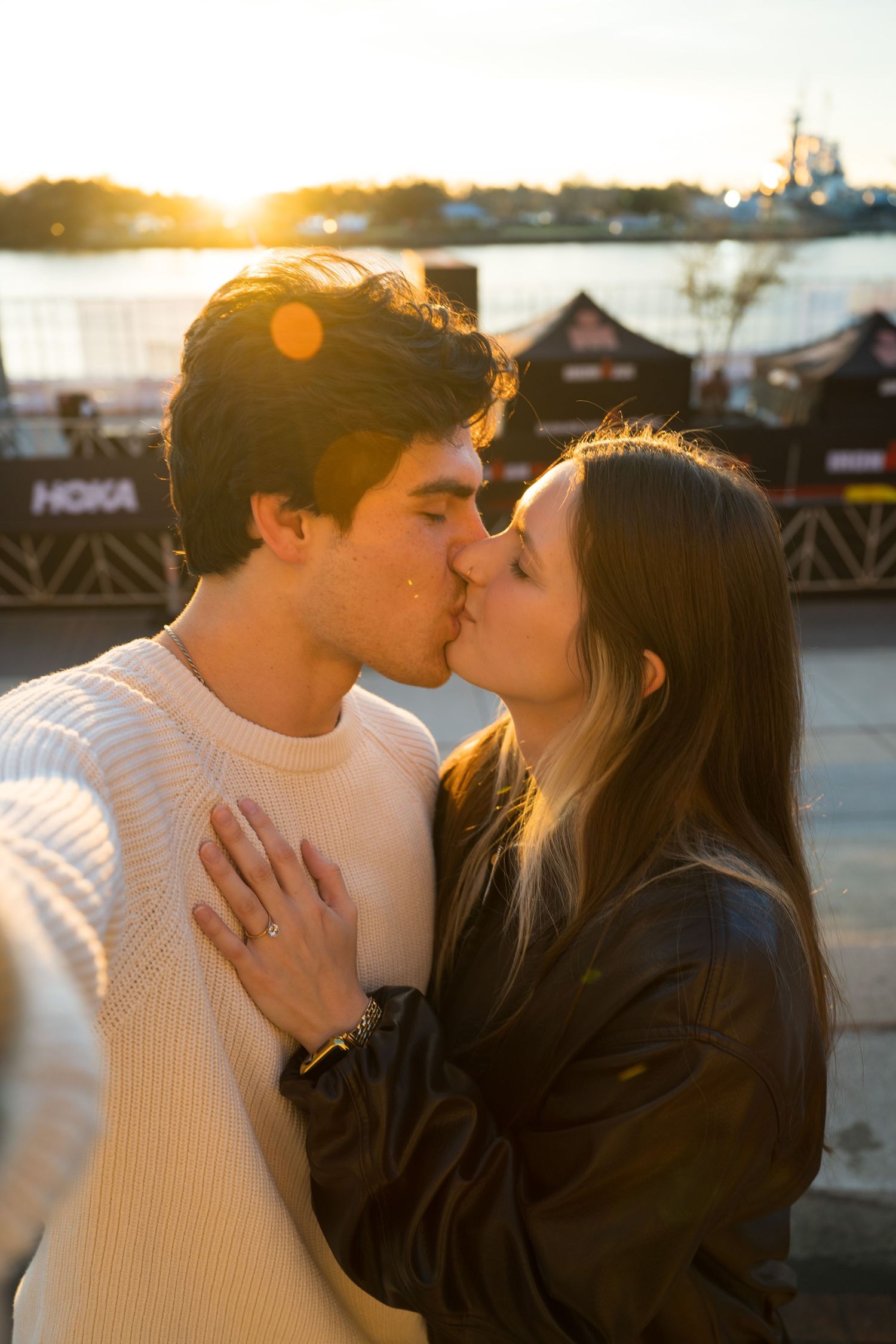 A romantic couple sharing a kiss at sunset by the waterfront with golden sunlight and buildings in the background.