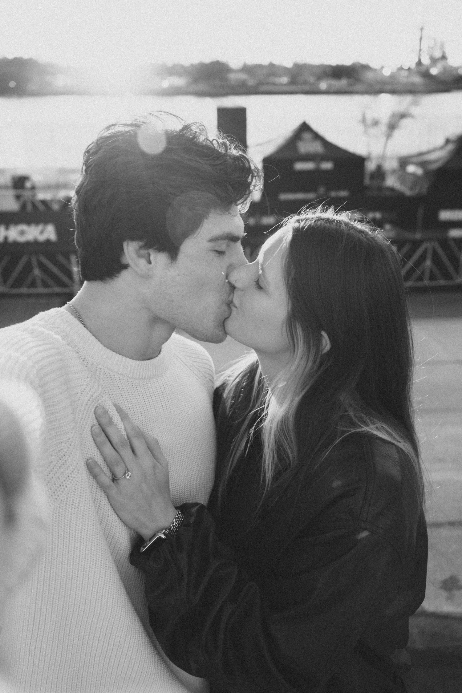 A black and white photo of a couple kissing outdoors with a waterfront setting in the background.