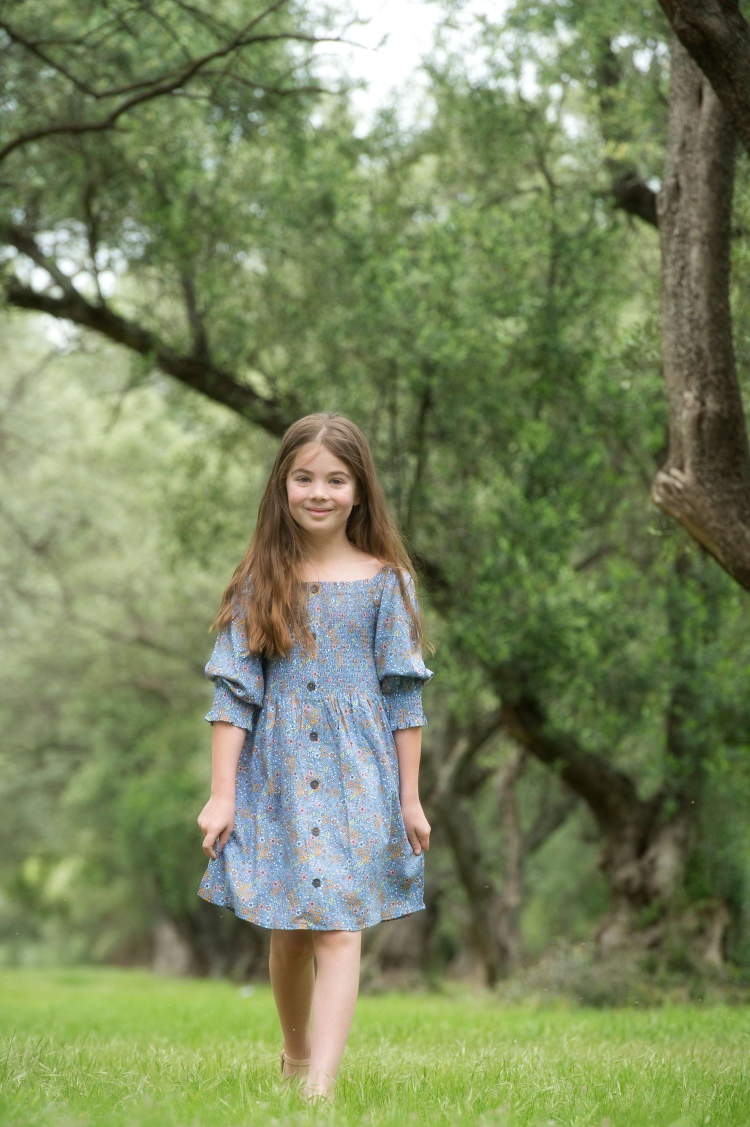 Young girl in blue button-front dress walking through a lush green garden setting.