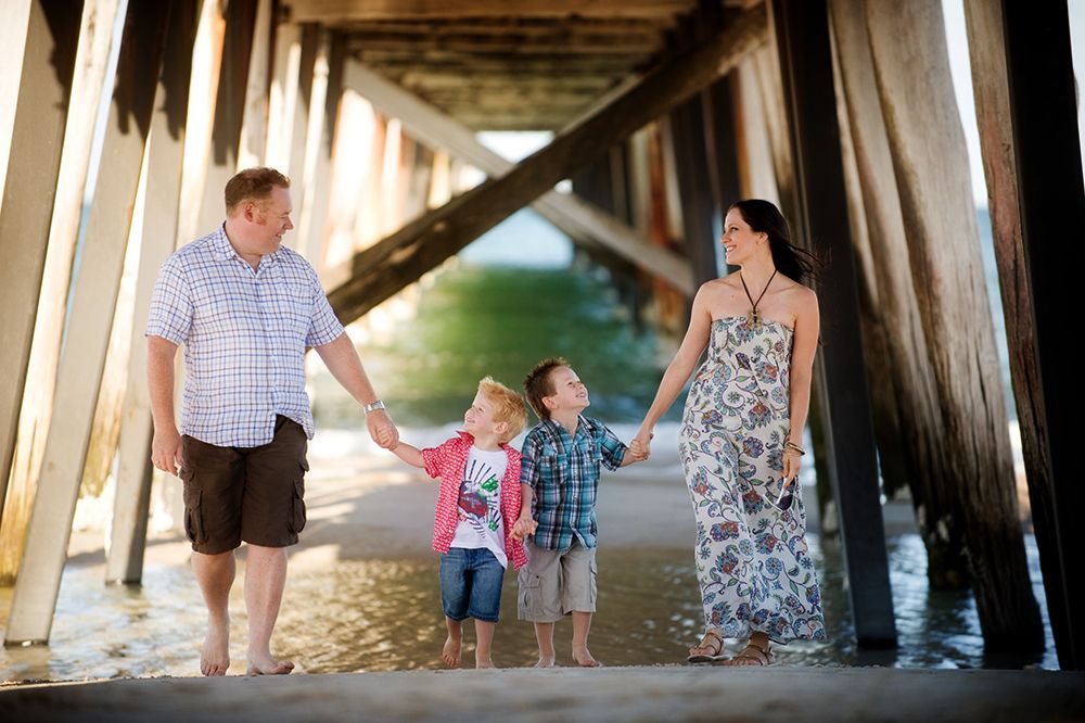 Family walking and posing under wooden pier structure at beach.