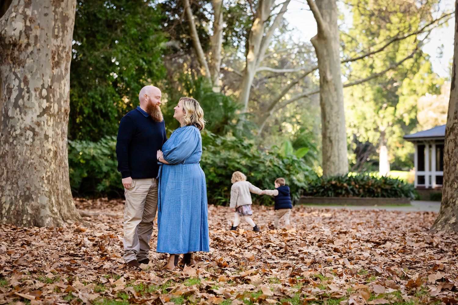 Family of four enjoying autumn leaves in a wooded park, parents watching toddlers play among fallen leaves.