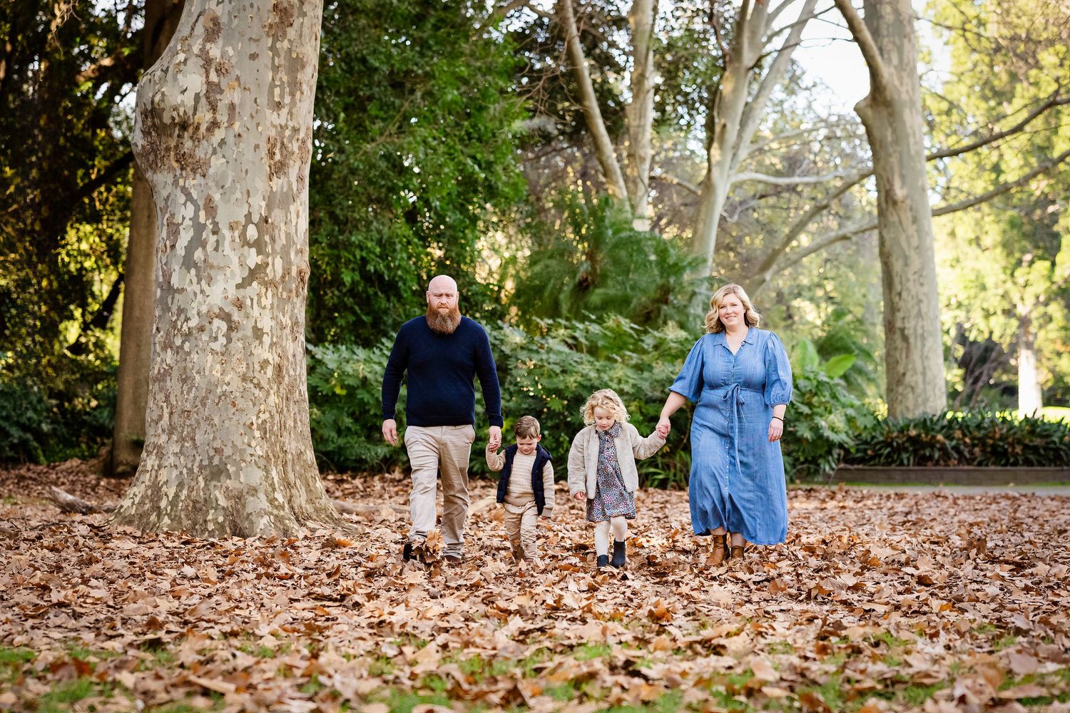 Family of five walking through autumn leaves in a tree-lined park, parents holding children's hands outdoors.