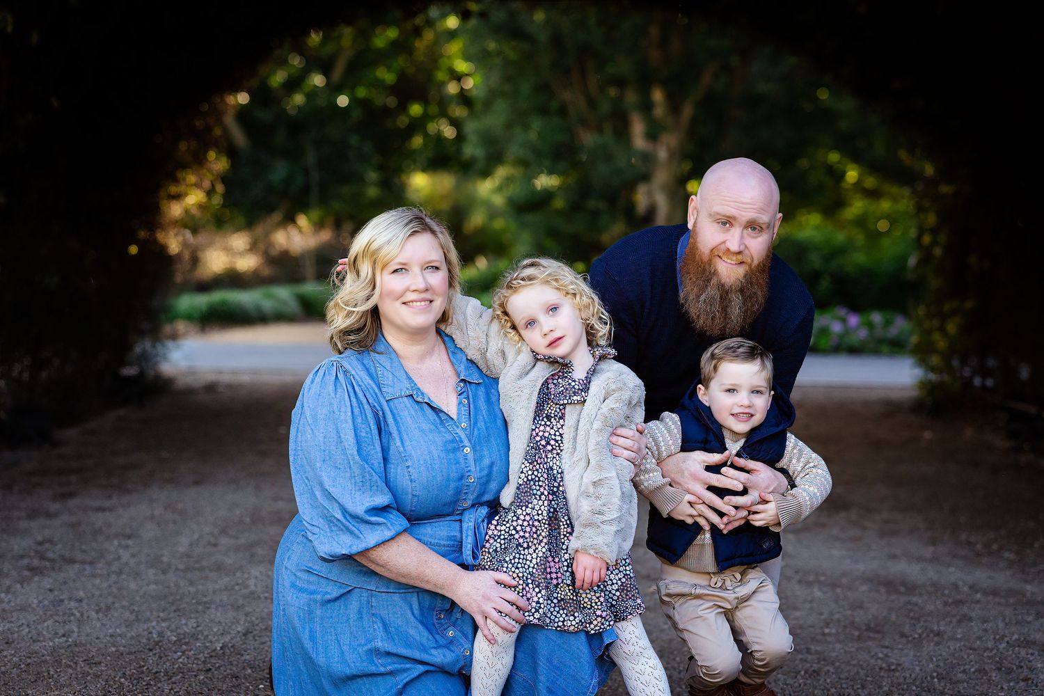 Family of four posing outdoors under arch with lush greenery, parents smiling with young daughter and son.