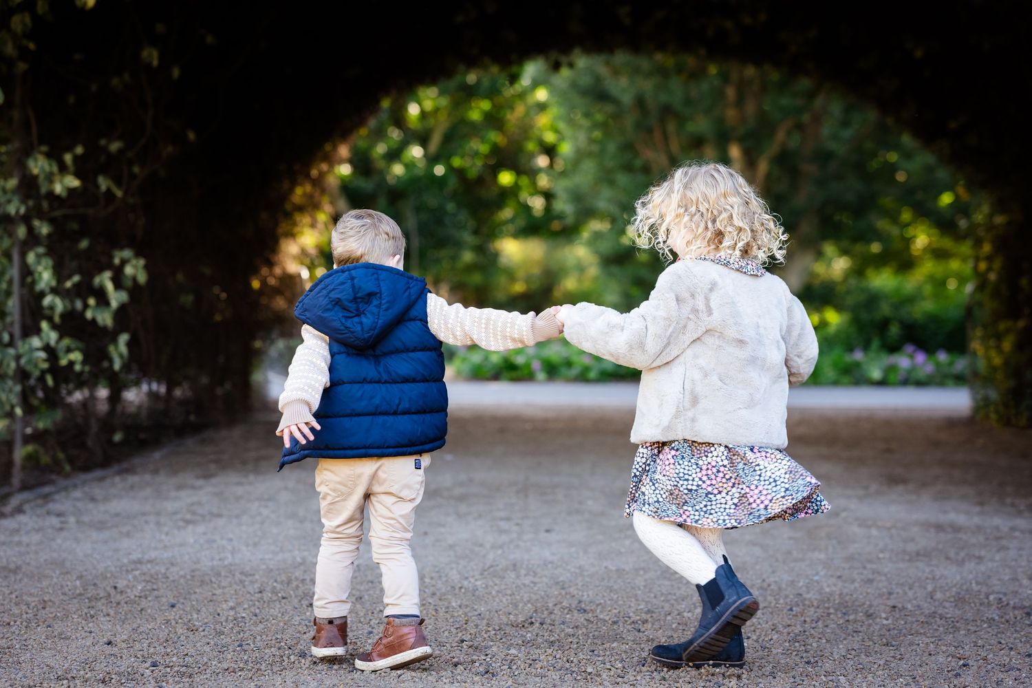 Two young children holding hands walking through a garden archway, boy in blue vest and girl in floral skirt.
