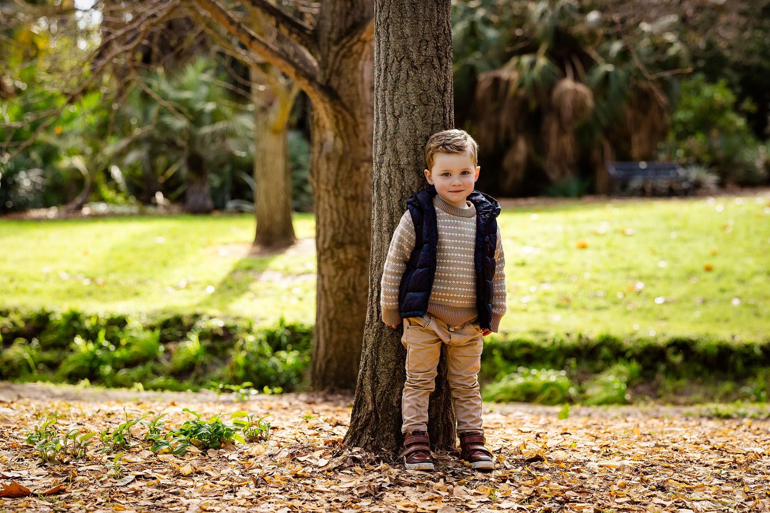 Young boy in vest and khakis leaning against a large tree trunk in an autumn park with fallen leaves.