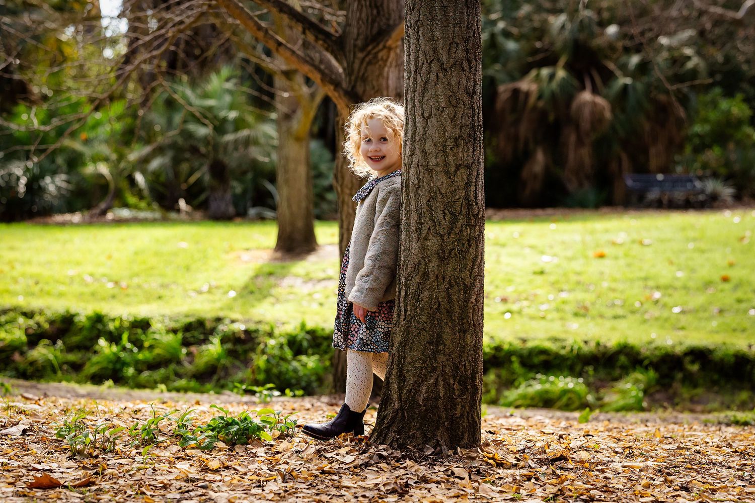 A young blonde child peeks around a tree trunk in an autumn park, wearing a grey dress and black boots.