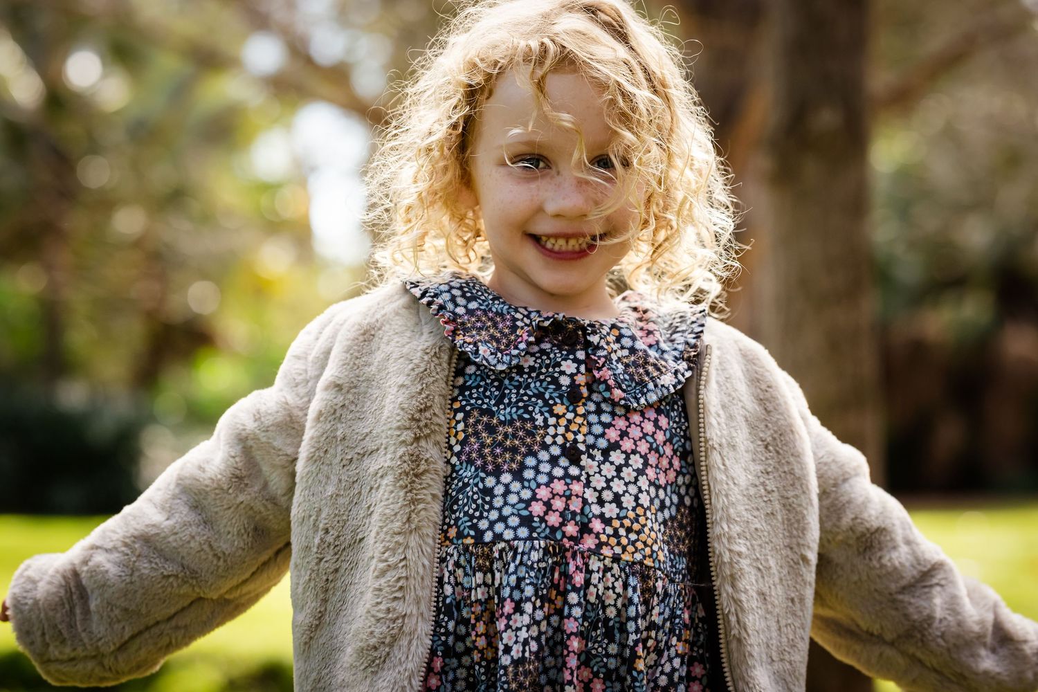 Smiling young girl with curly blonde hair wearing a floral dress and fuzzy beige jacket outdoors.