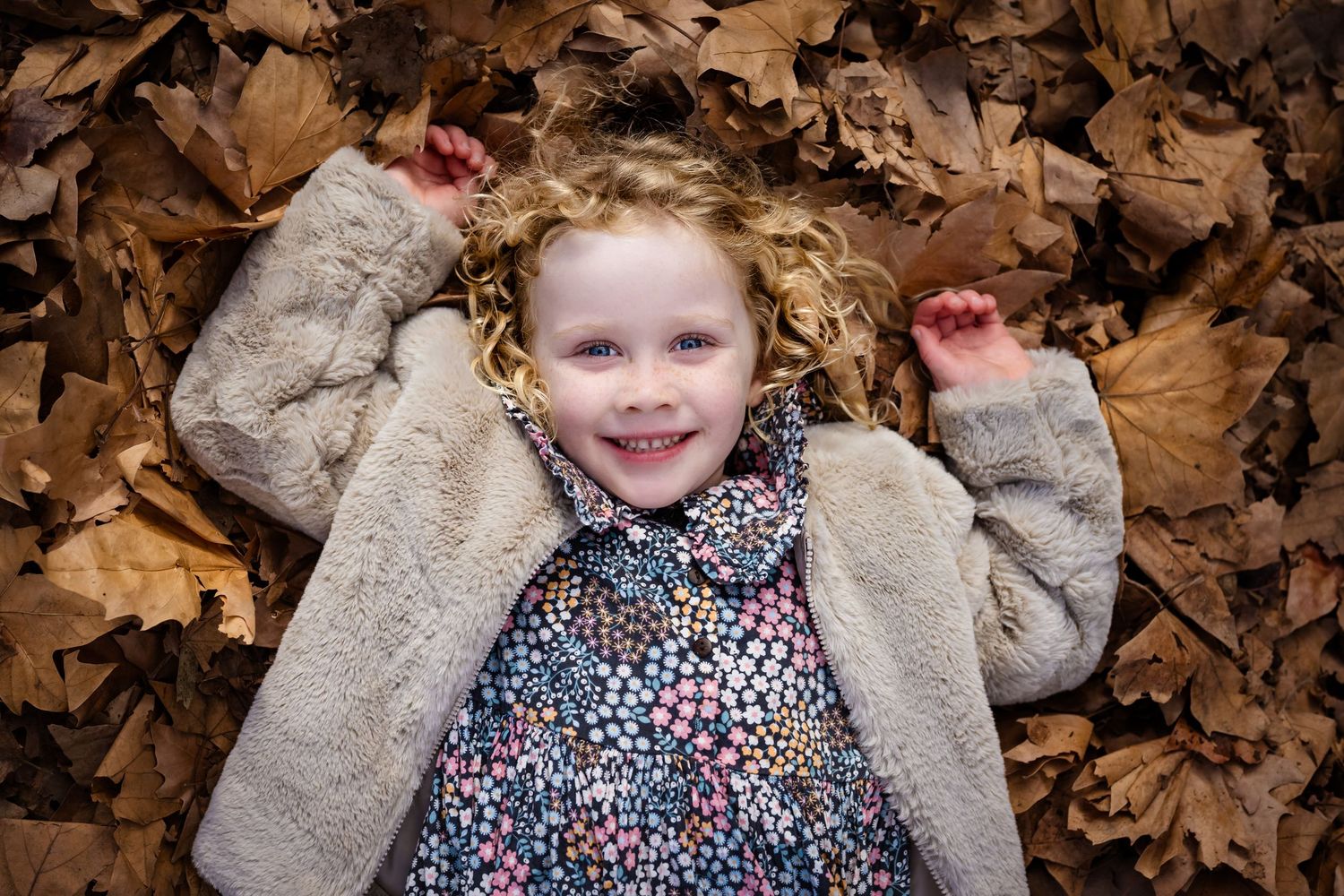 Happy young girl with curly blonde hair lying in autumn leaves wearing a floral dress and beige jacket.