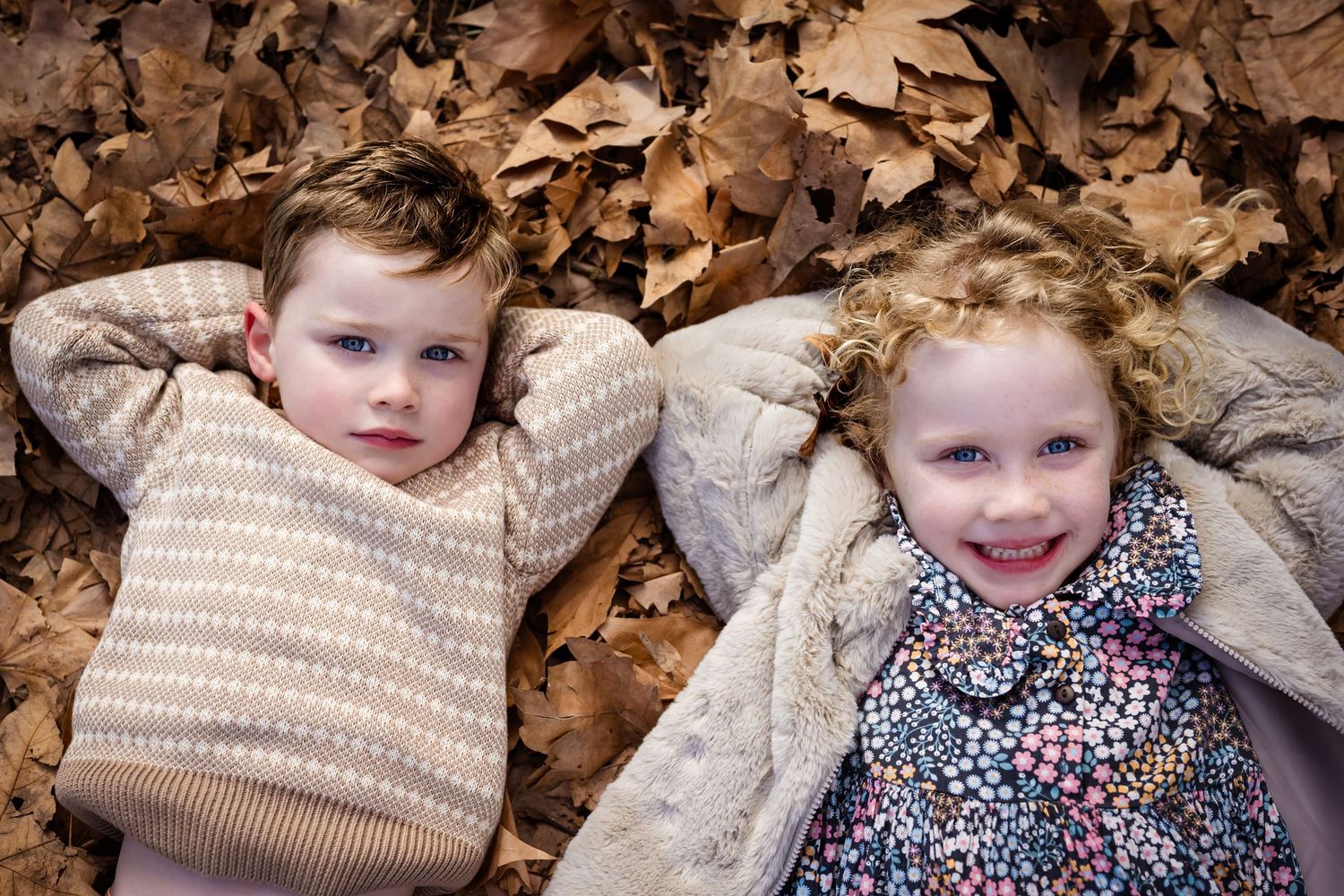 Two young children lie smiling in a bed of autumn leaves, wearing cozy sweaters in a fall outdoor setting.