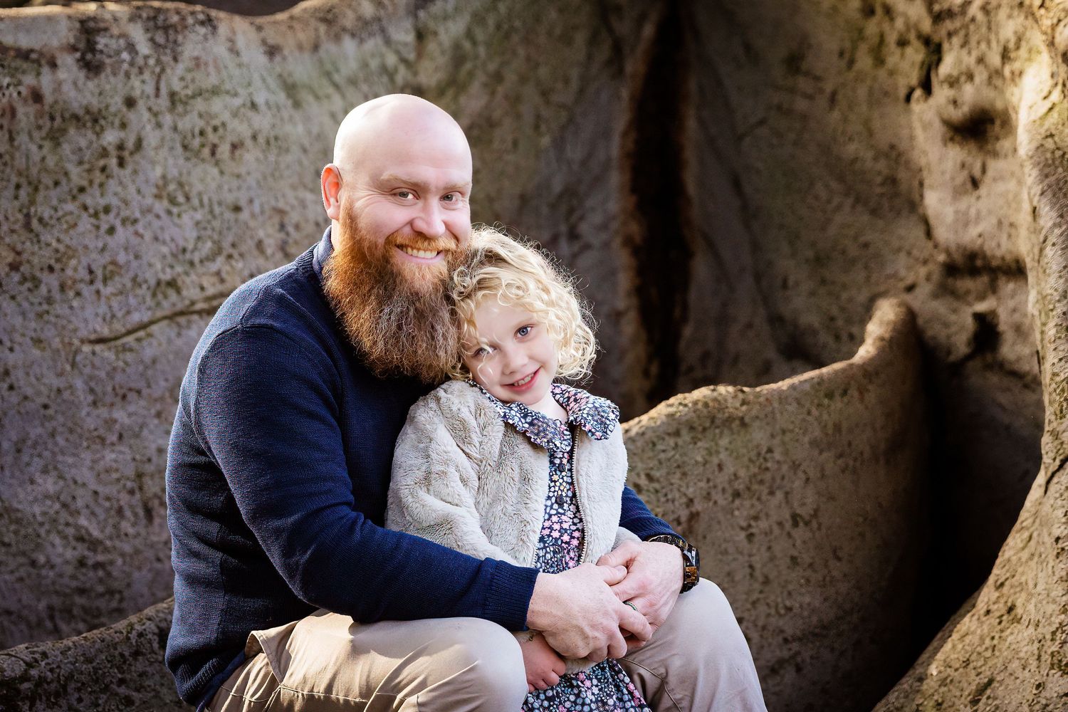 Bald bearded man sitting with young girl in floral dress inside a large tree hollow, smiling warmly.