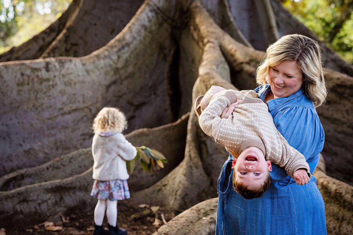Mother holds toddler upside down laughing near large tree roots while young girl stands nearby.
