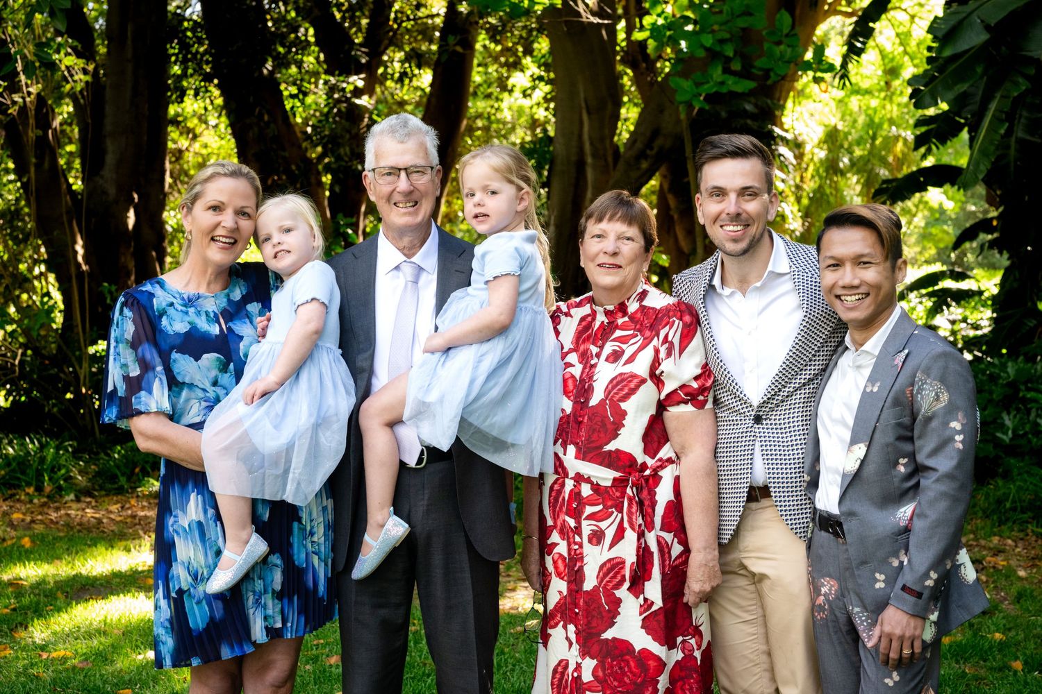 Multi-generational family portrait outdoors, smiling group of seven including children and grandparents in a lush green garden.