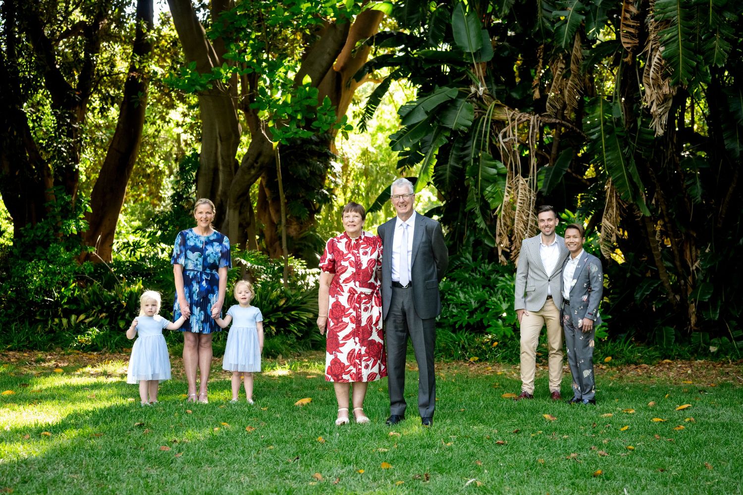 Family group portrait on green lawn with lush tropical trees, featuring adults and children in formal attire.