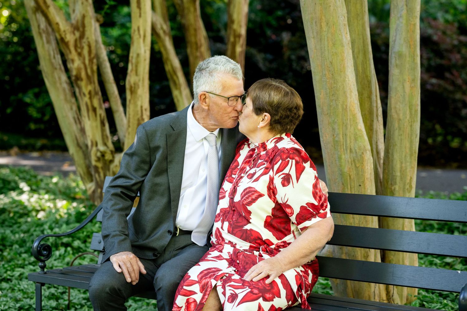 Elderly couple sharing a romantic kiss on a park bench surrounded by lush green trees.