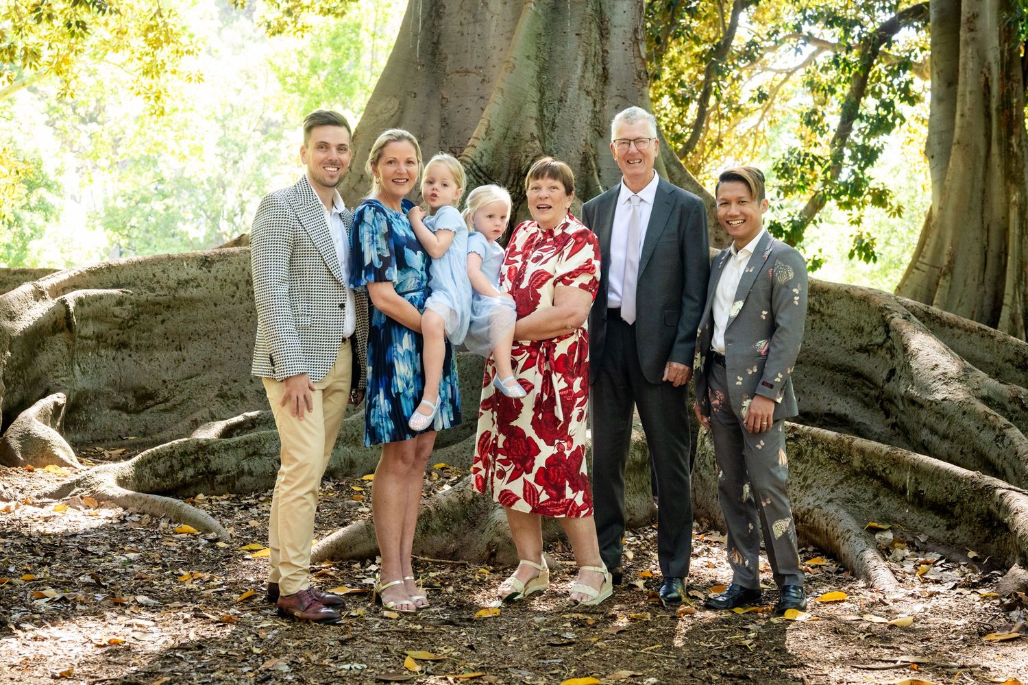 Multi-generational family group smiling together outdoors near large tree roots in a lush garden setting.