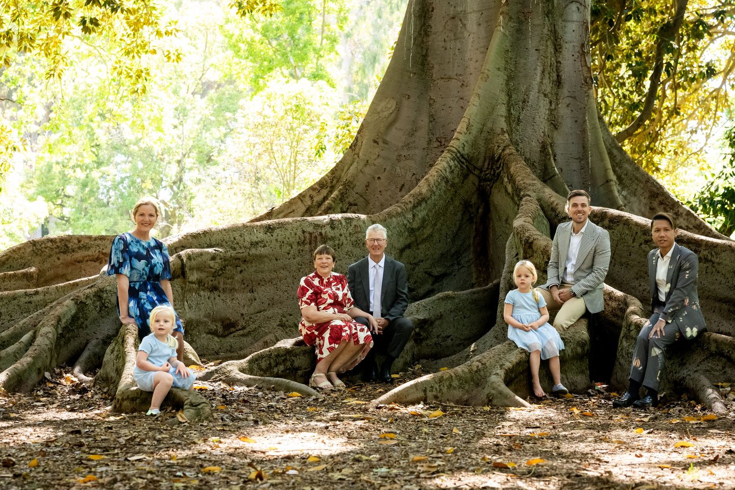 Multi-generational family portrait posed around large tree roots in a lush green outdoor setting.