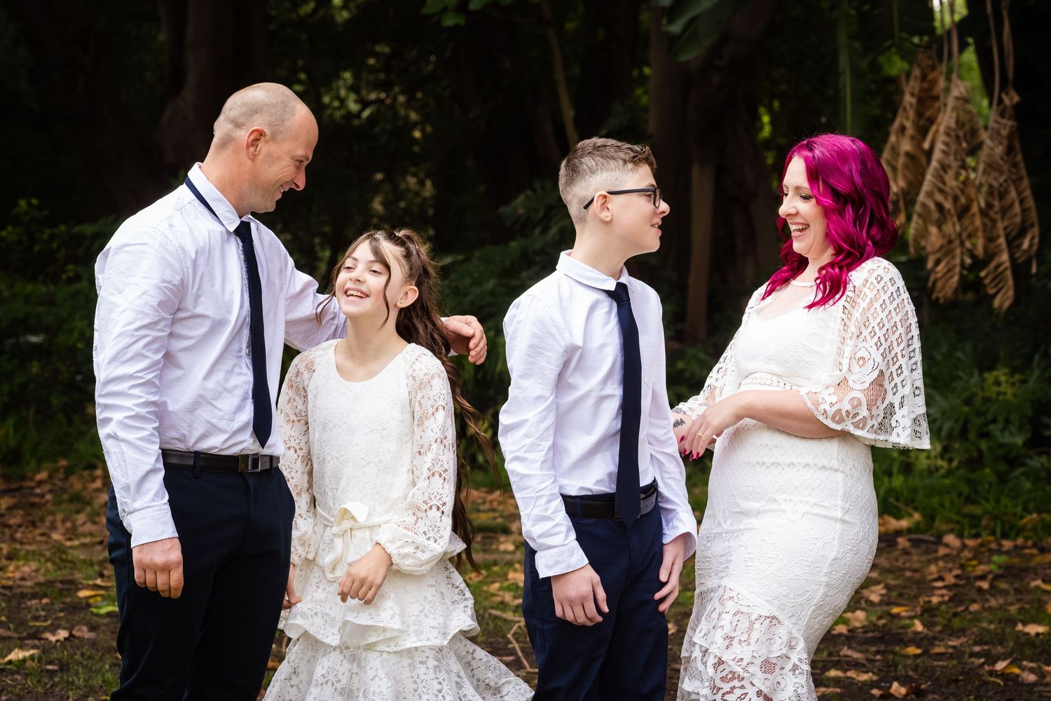 Family of four in white formal attire posing outdoors, parents and two children smiling together in a garden setting.