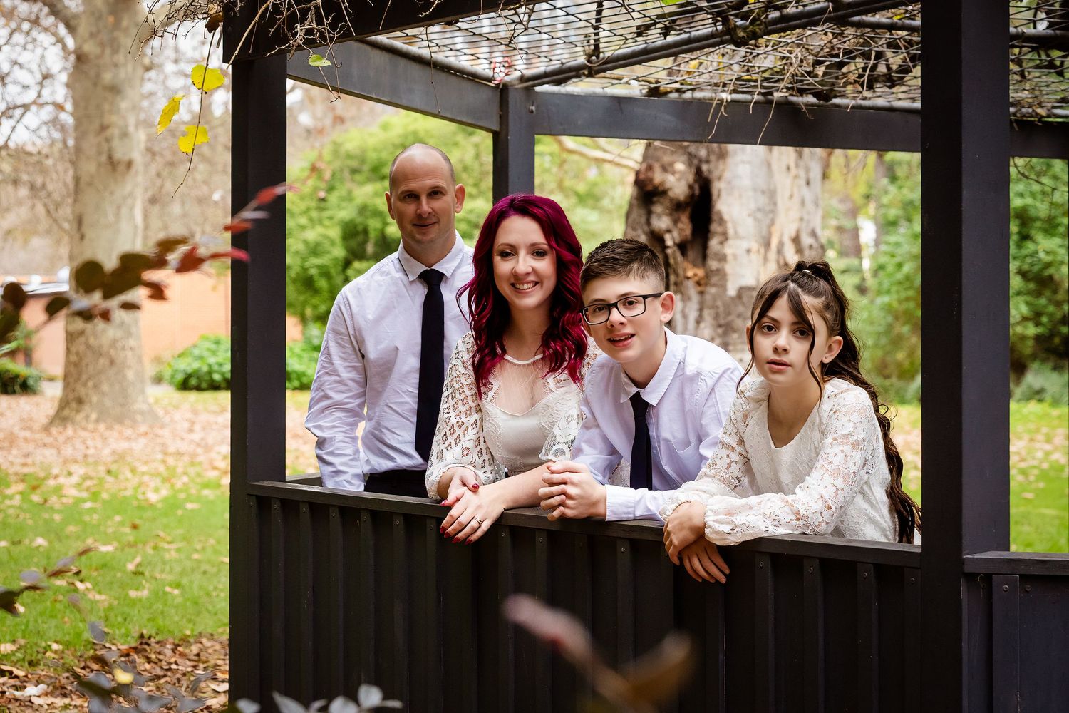 Family of four posing under a dark pergola in a garden, dressed in formal attire for a wedding celebration.