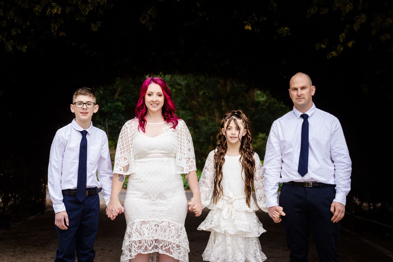 Family of four holding hands outdoors, bride in lace dress with red hair, groom bald in white shirt, two children dressed formally.