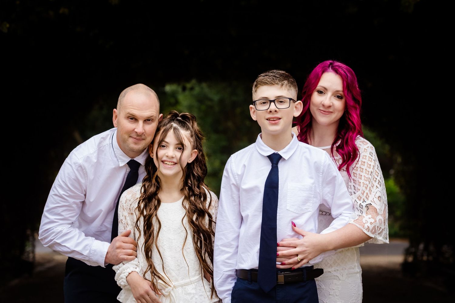Family of four posing outdoors, parents with two children dressed formally, woman with pink hair, boy in tie, girl in white dress.