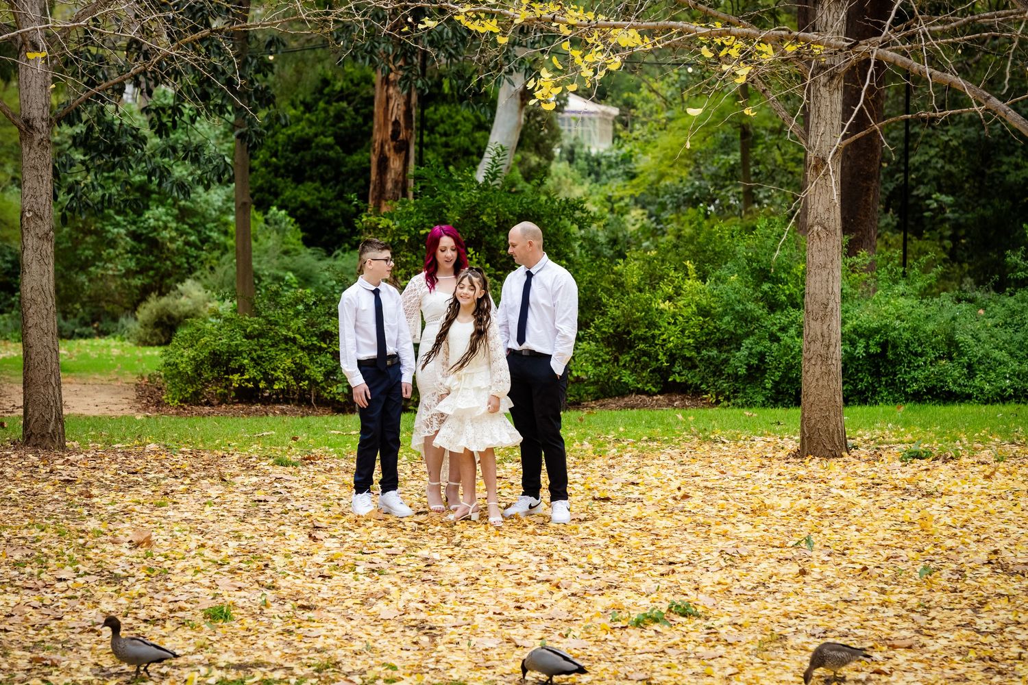 Family of three posing among autumn leaves in a park, with geese nearby and lush green trees in the background.