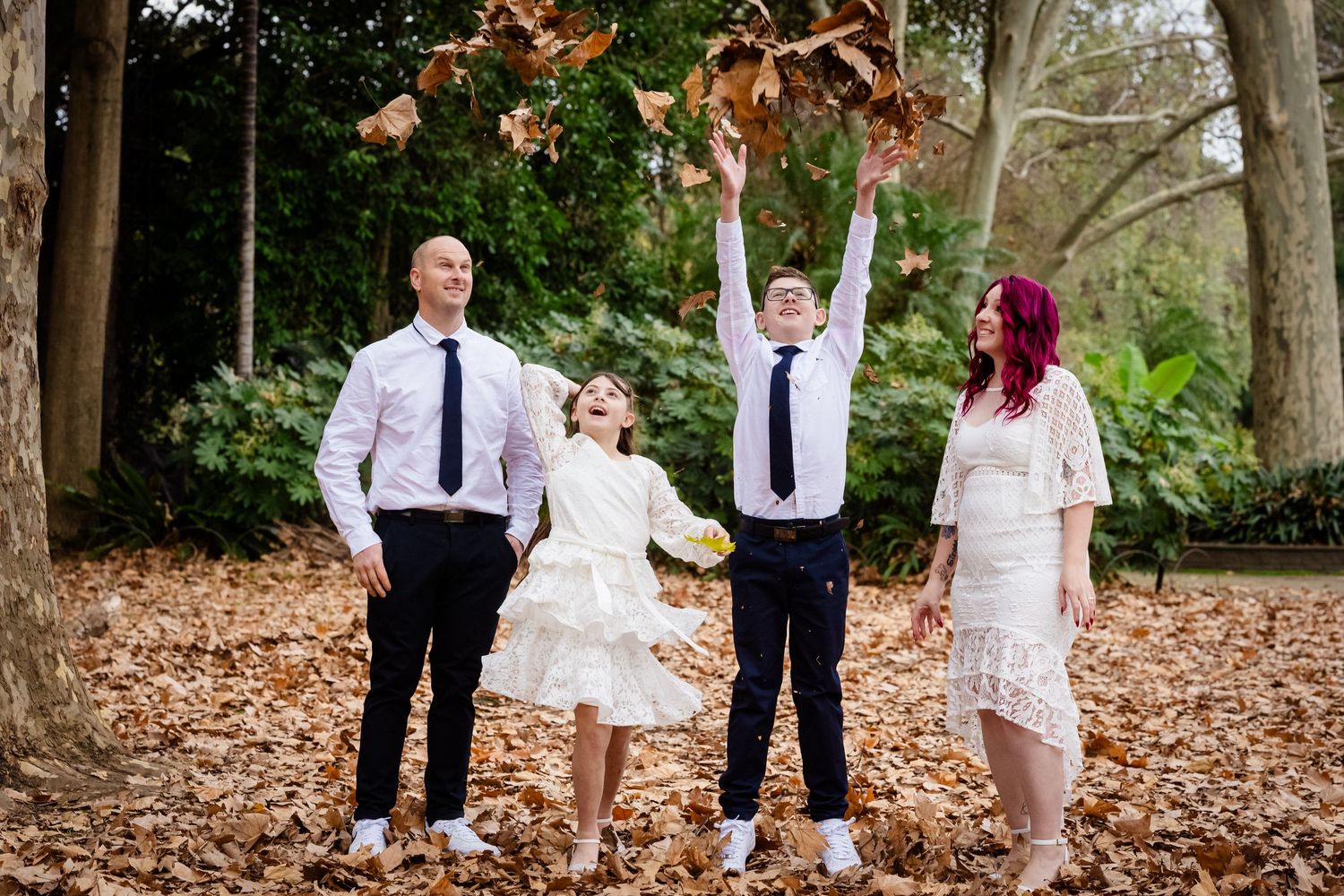 Family of four enjoying autumn leaves in a park, children tossing leaves in the air, dressed in formal white and navy attire.