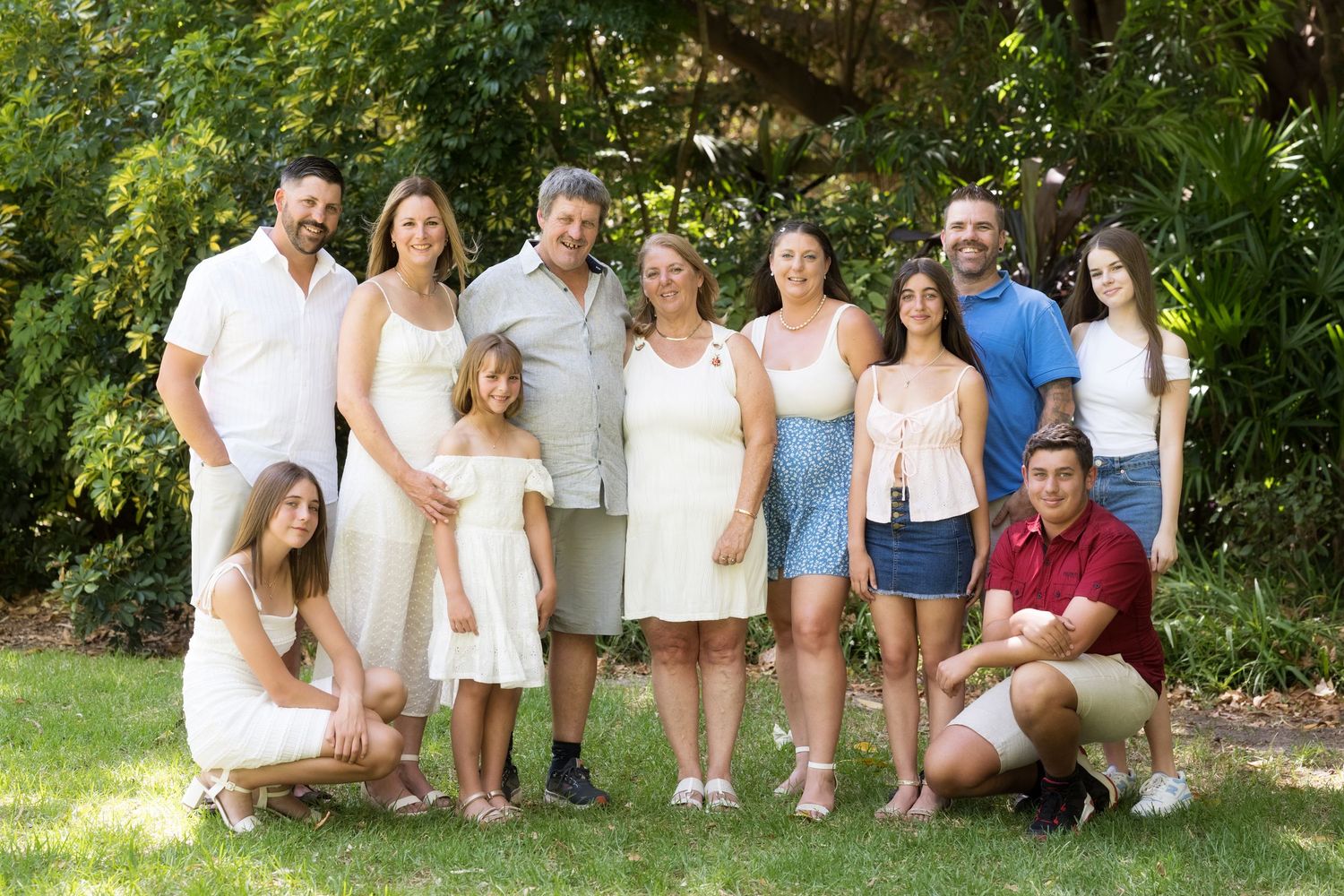 Multi-generational family group poses together outdoors on a sunny day in casual summer attire.