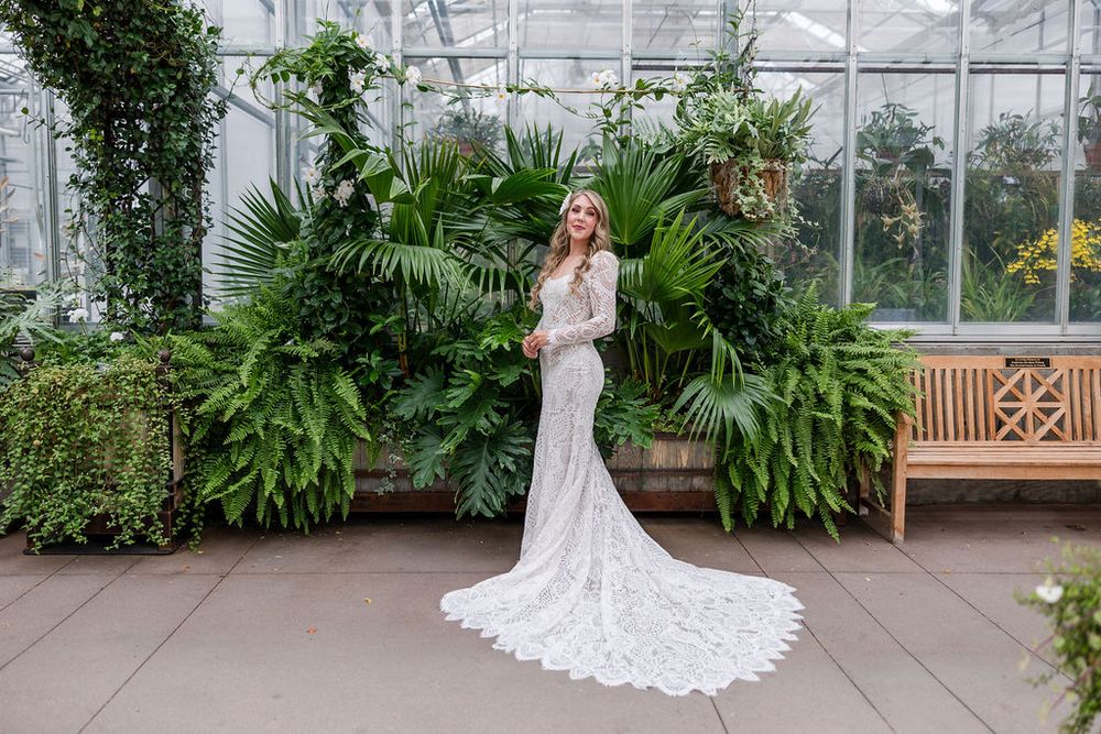 A bride in a white lace wedding dress poses in a greenhouse filled with lush tropical plants and greenery.