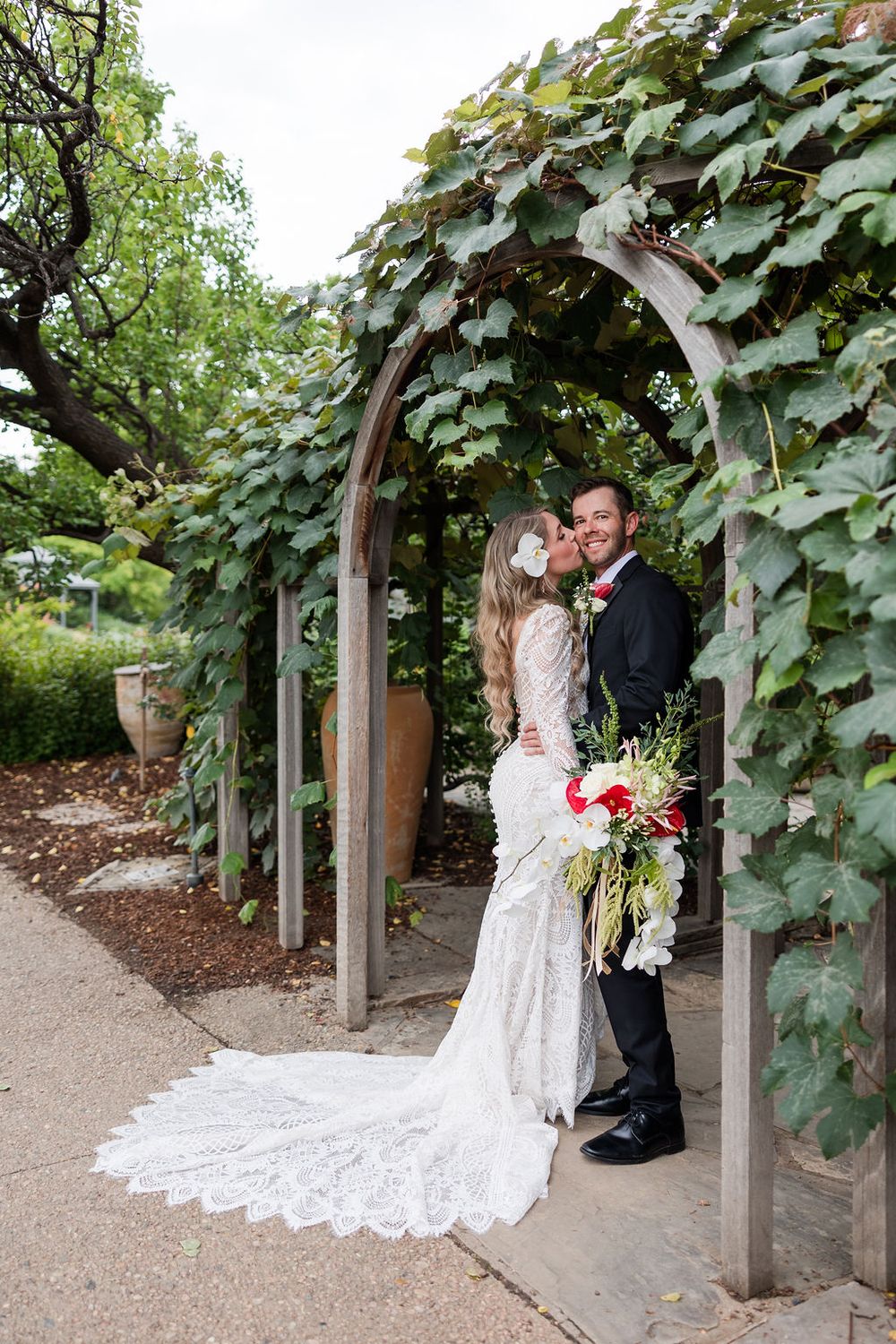 A couple shares a romantic moment under an ivy-covered garden archway, with a stunning lace wedding dress trailing behind.