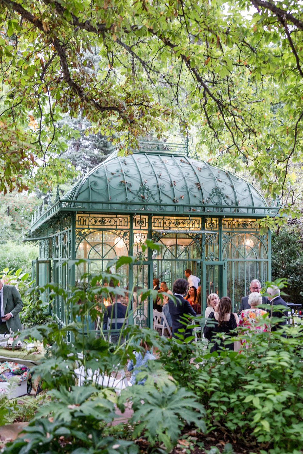 A vintage greenhouse with glass panels and ornate metalwork surrounded by lush green foliage and garden visitors.