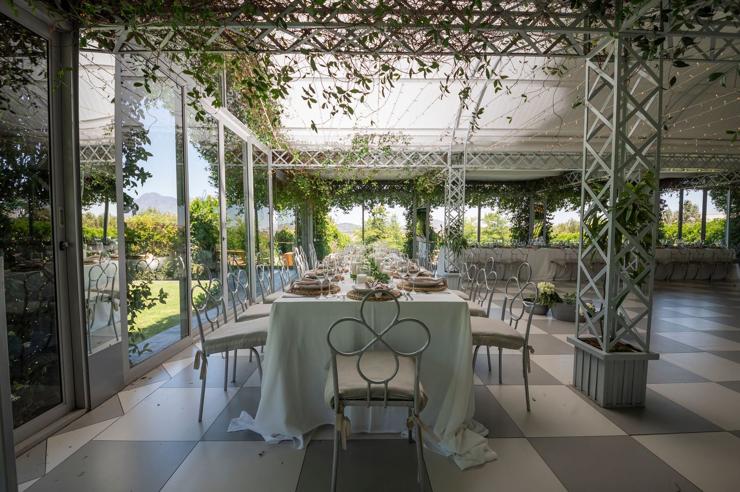 A long covered walkway with white metal lattice work and hanging plants stretches through a garden setting.