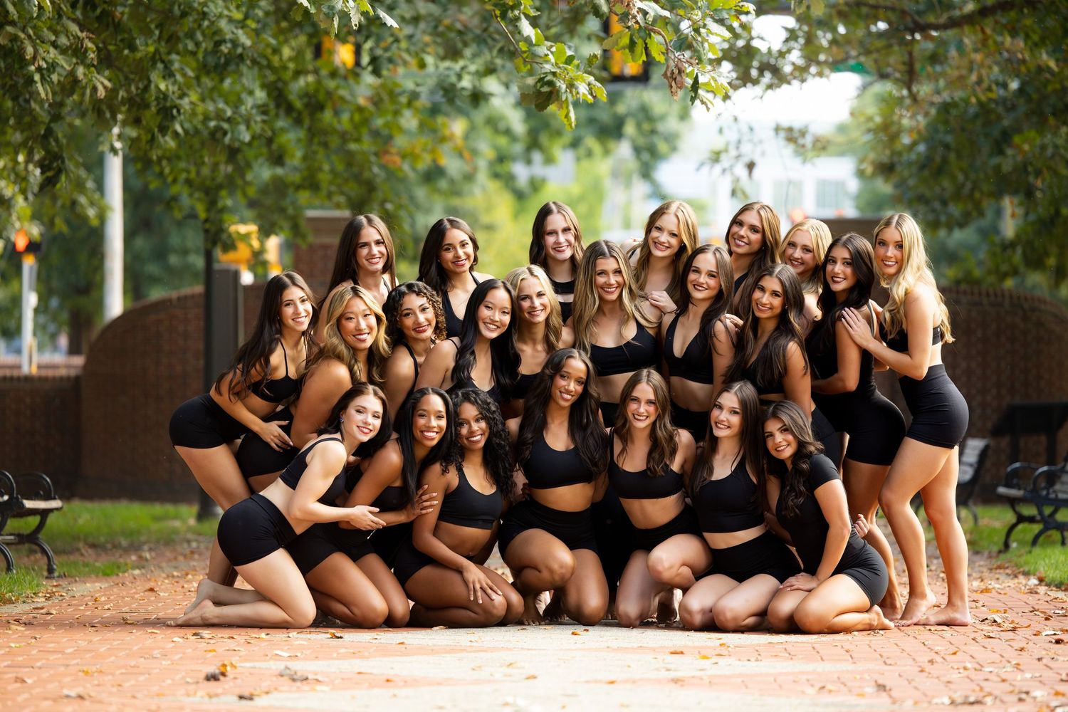 Group of dancers in black outfits pose together outdoors for a team photo on a brick path.