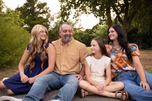 A family group sits casually together outdoors on grass during a sunny day for a relaxed portrait session.